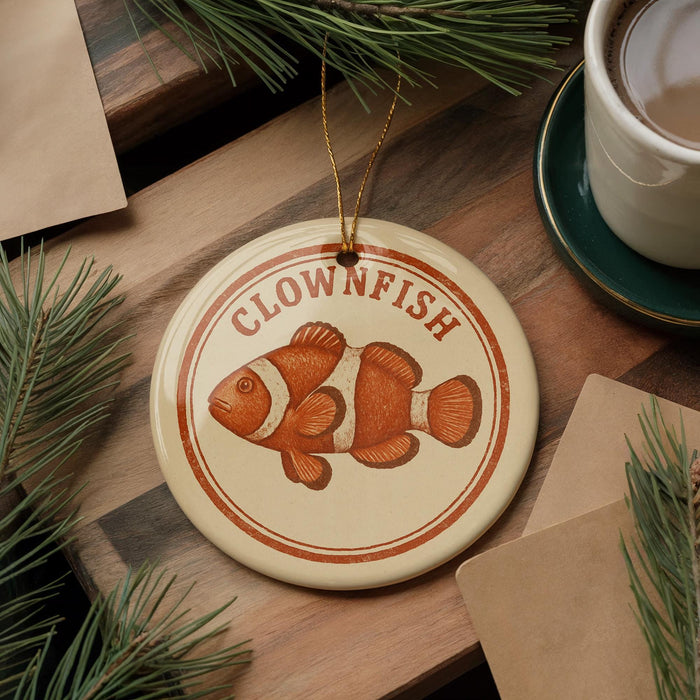 A round ornament featuring an orange clownfish is displayed on a wooden surface, surrounded by pine branches and a cup of coffee.