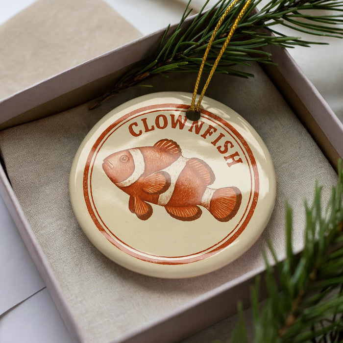 A round ornament with a clownfish design is displayed in a box, surrounded by pine branches.