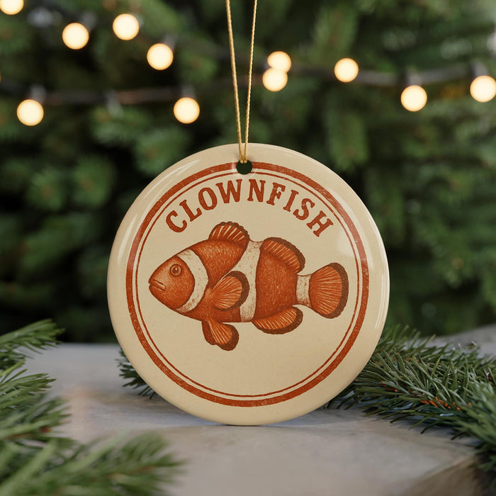 A round ornament with a clownfish design, hanging from a string and placed on a surface with pine branches in the background.