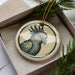 A ceramic ornament in the shape of a parrot is displayed in a box, surrounded by pine branches.