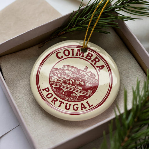 A circular ornament with the text "COIMBRA PORTUGAL" printed on it, placed inside a box with pine branches.