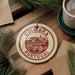 A circular ornament with the text "COIMBRA PORTUGAL" is displayed on a wooden surface, surrounded by pine branches and a cup of coffee.