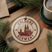 A round ornament with the text "COLOGNE GERMANY" and an illustration of a cathedral, placed on a wooden surface next to a cup of coffee and some pine branches.