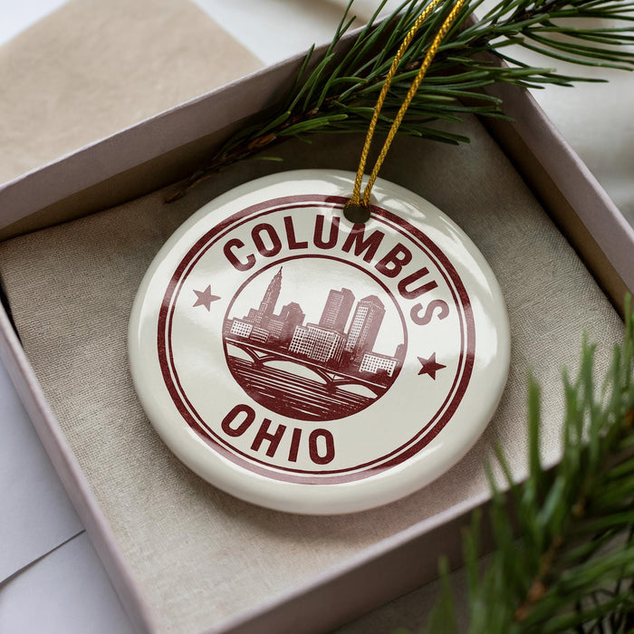 A white ornament with the text "COLUMBUS OHIO" printed on it, placed in a box with pine branches.