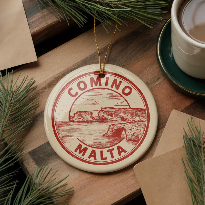A circular ornament with the text "COMINO MALTA" is displayed on a wooden surface, surrounded by pine branches and a cup of coffee.