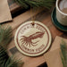 a round ornament with a picture of a squid and the text "Common squid" on it, placed on a wooden surface. There is also a cup of coffee and some pine branches in the background.