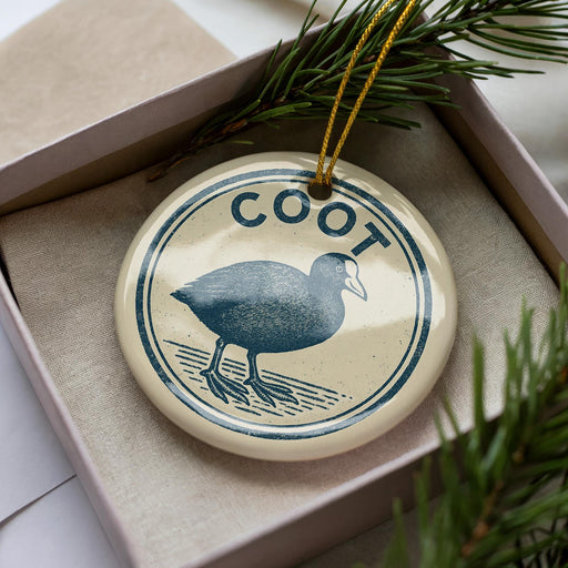 A ceramic ornament in a box, featuring a blue bird design with the word "Coot" written around it.