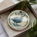 A ceramic ornament in a box, featuring a blue bird design with the word "Coot" written around it.