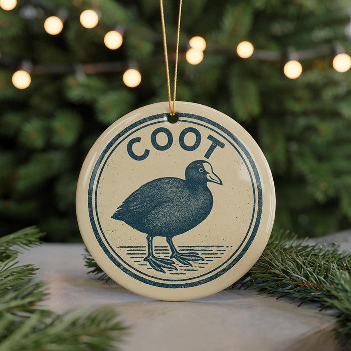 A round ornament with the word "Coot" on it, featuring a blue illustration of a bird, is hanging from a string in front of a Christmas tree with twinkling lights.