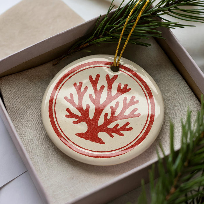 A round ornament with a red tree design is displayed in a box, surrounded by pine branches.