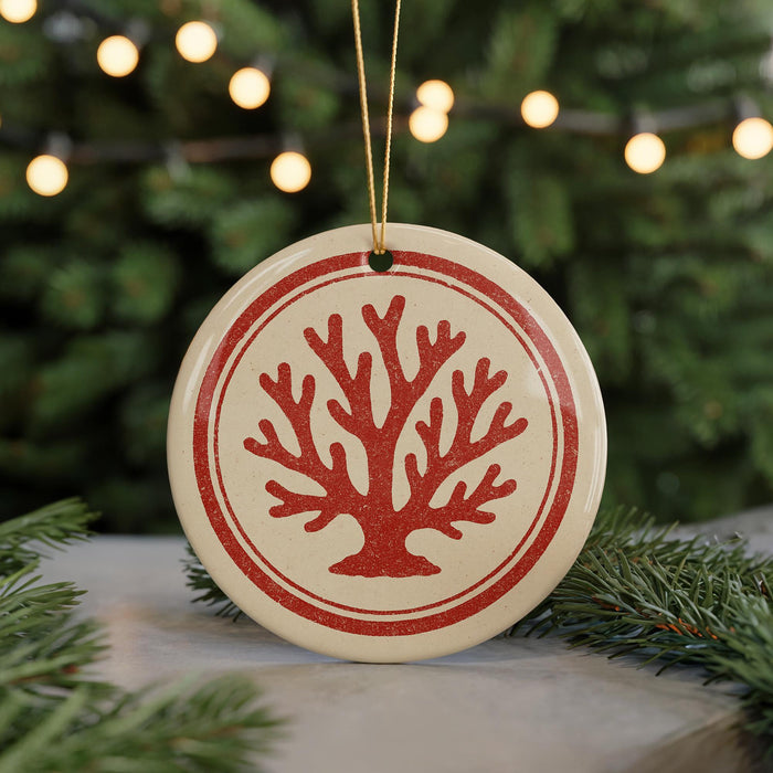 A round ornament with a red tree design is hanging from a string, surrounded by pine branches and twinkling lights in the background.