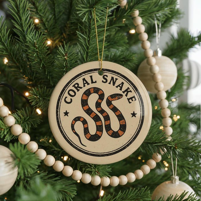 A round ornament with a black and orange snake design hangs from a Christmas tree, surrounded by white beads and greenery.
