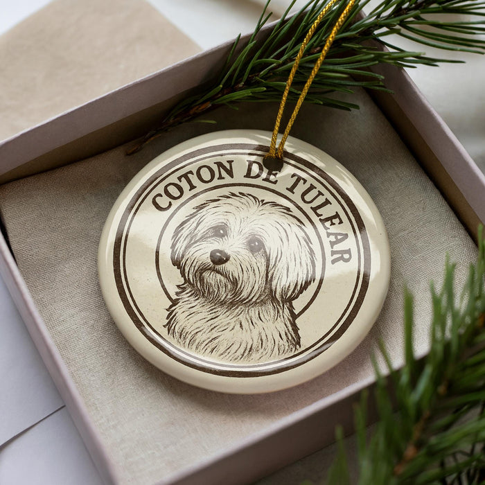 A circular ornament with a dog image and the text "COTON DE TULFAR" printed on it, placed inside a box with pine branches.