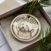 A circular ornament with a dog image and the text "COTON DE TULFAR" printed on it, placed inside a box with pine branches.