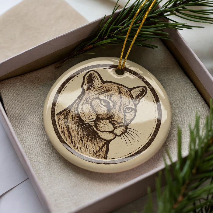 A ceramic ornament in the shape of a cat's face, with a brown and beige color scheme, is displayed in a box surrounded by pine branches.