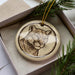 A ceramic ornament in the shape of a cat's face, with a brown and beige color scheme, is displayed in a box surrounded by pine branches.