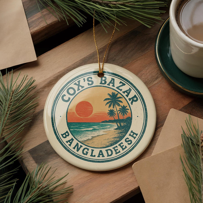 A circular ornament with the text "Cox's Bazar Bangladesh" and a tropical beach scene, hanging on a wooden surface with pine branches and a cup of coffee nearby.