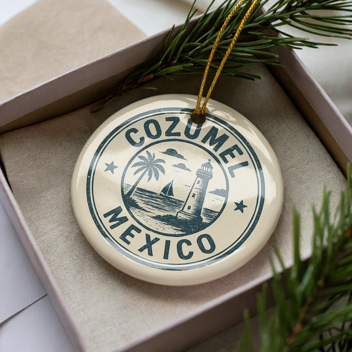 A decorative ornament with the text "COZUMEL MEXICO" printed on it, surrounded by a lighthouse and palm trees, is displayed in a box with pine branches.