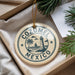 A circular ornament with the text "COZUMEL MEXICO" printed on it, surrounded by a lighthouse and palm trees, is displayed in a box with pine branches.