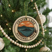 A circular ornament on a Christmas tree, featuring a scenic landscape with mountains and a lake, with the text "Crater Lake National Park" prominently displayed.