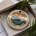 A circular ornament with the text "CRATER LAKE NATIONAL PARK" printed on it, surrounded by pine branches and pine needles.