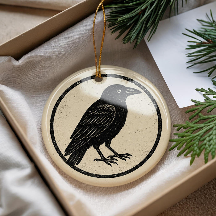 A round ornament featuring a black bird is displayed in a box, surrounded by pine branches.
