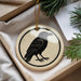 A round ornament featuring a black bird is displayed in a box, surrounded by pine branches.