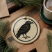 a ceramic ornament with a black bird design, placed on a wooden surface next to a cup of coffee and some pine branches.