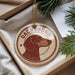 A round ornament with a dachshund dog's head design, hanging from a string, placed inside a box with pine branches.