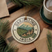 A decorative ornament with the text "DARJEELING INDIA" and a landscape image, placed on a wooden surface next to a cup of coffee and some pine branches.