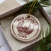 A decorative ornament in a box, featuring a circular design with a city skyline and the text "DES MOINES IOWA" printed on it.