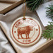 A decorative ornament in the shape of a cow, with the text "Domestic Cow" printed on it, is displayed in a box with pine branches.
