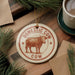 a decorative ornament with the text "Domestic Cow" and a cow illustration, placed on a wooden surface alongside a cup of coffee and some pine branches.