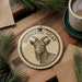 a round ornament with a goat's head on it, placed on a wooden surface next to a cup of coffee and some pine branches.