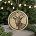 A round ornament with a goat's head and horns, surrounded by a decorative border, is displayed in front of a Christmas tree with twinkling lights.