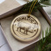 A decorative ornament featuring a pig illustration, placed in a box with pine branches.