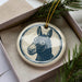 A ceramic ornament in the shape of a donkey's head, with a blue and white design, is displayed in a box with pine branches.