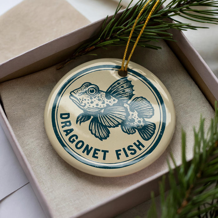 A round ornament with a blue and white design featuring a fish, placed inside a box with pine branches.