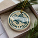 A round ornament with a blue and white design featuring a fish, placed inside a box with pine branches.