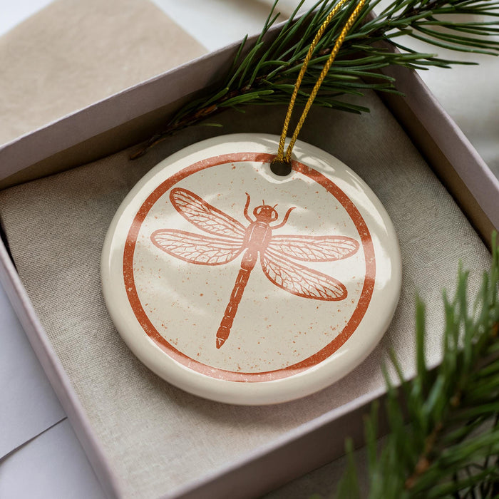 A ceramic ornament in the shape of a dragonfly is displayed in a box, surrounded by pine branches.