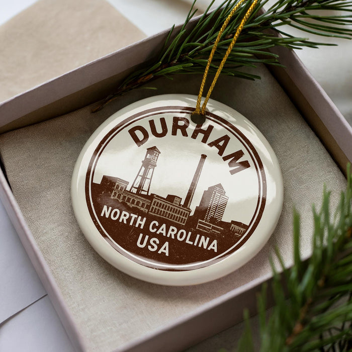 A decorative ornament with the text "DURHAM NORTH CAROLINA USA" on it, placed in a box with pine branches.