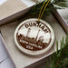 A decorative ornament with the text "DURHAM NORTH CAROLINA USA" on it, placed in a box with pine branches.