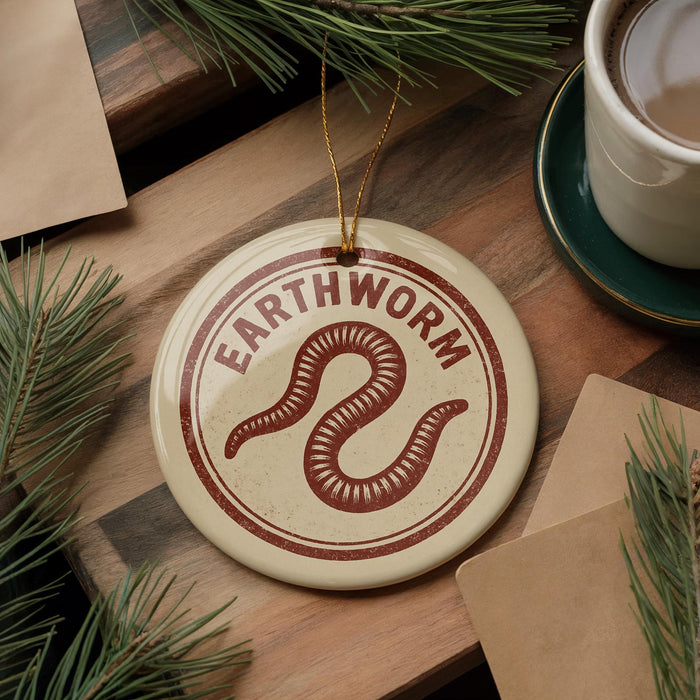 a circular ornament with the word "Earthworm" written on it, placed on a wooden surface next to a cup of coffee and some pine branches.