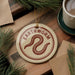 a circular ornament with the word "Earthworm" written on it, placed on a wooden surface next to a cup of coffee and some pine branches.