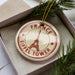 A circular ornament with the Eiffel Tower image and the text "FRANCE EIFEL Tower" printed on it, placed inside a box with pine branches.
