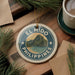 A circular ornament with the text "EL NIDO PHILIPPINES" and a scenic mountain landscape is displayed on a wooden surface, accompanied by a cup of coffee and pine branches.