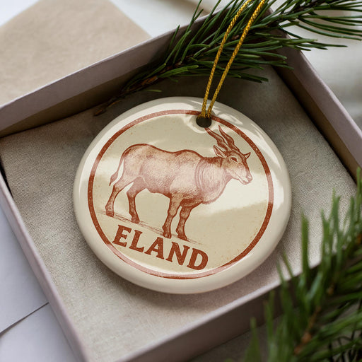 A circular ornament with a brown goat illustration and the word "ELAND" written on it, placed inside a box with pine branches.