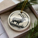 A ceramic ornament in the shape of a deer, with antlers, is displayed in a box with pine branches.