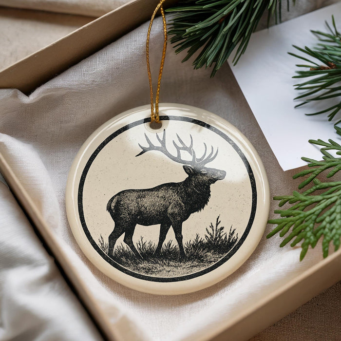 A decorative ornament featuring a deer standing in a grassy field, with a pine tree branch visible in the background.