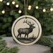 A ceramic ornament featuring a deer silhouette is displayed against a blurred background of a Christmas tree with twinkling lights.